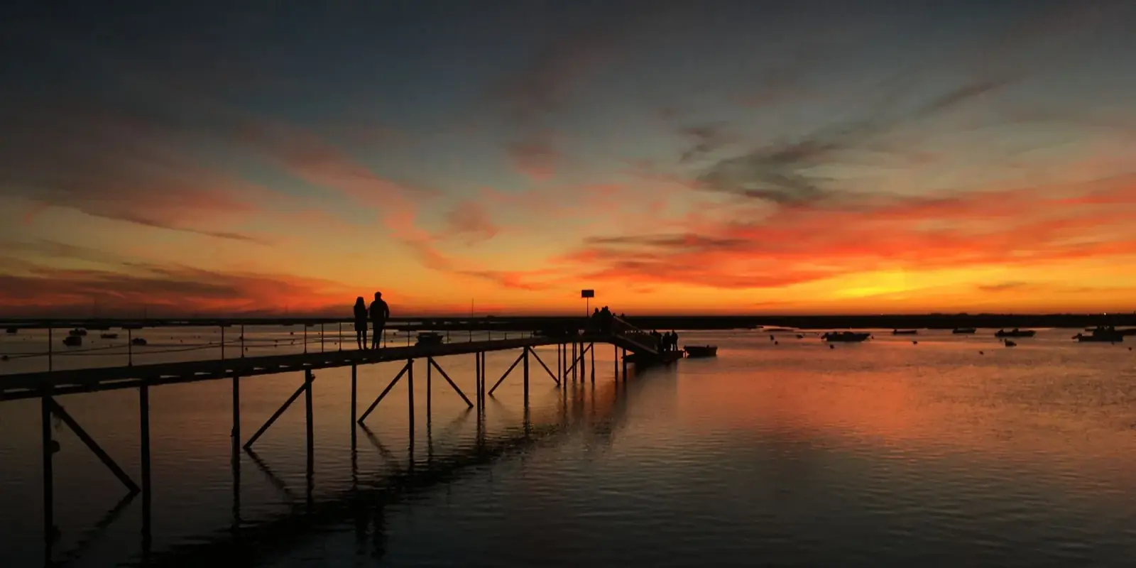 Cielo infuocato riflesso nella laguna di Faro, un pontile di legno e due sagome lontane