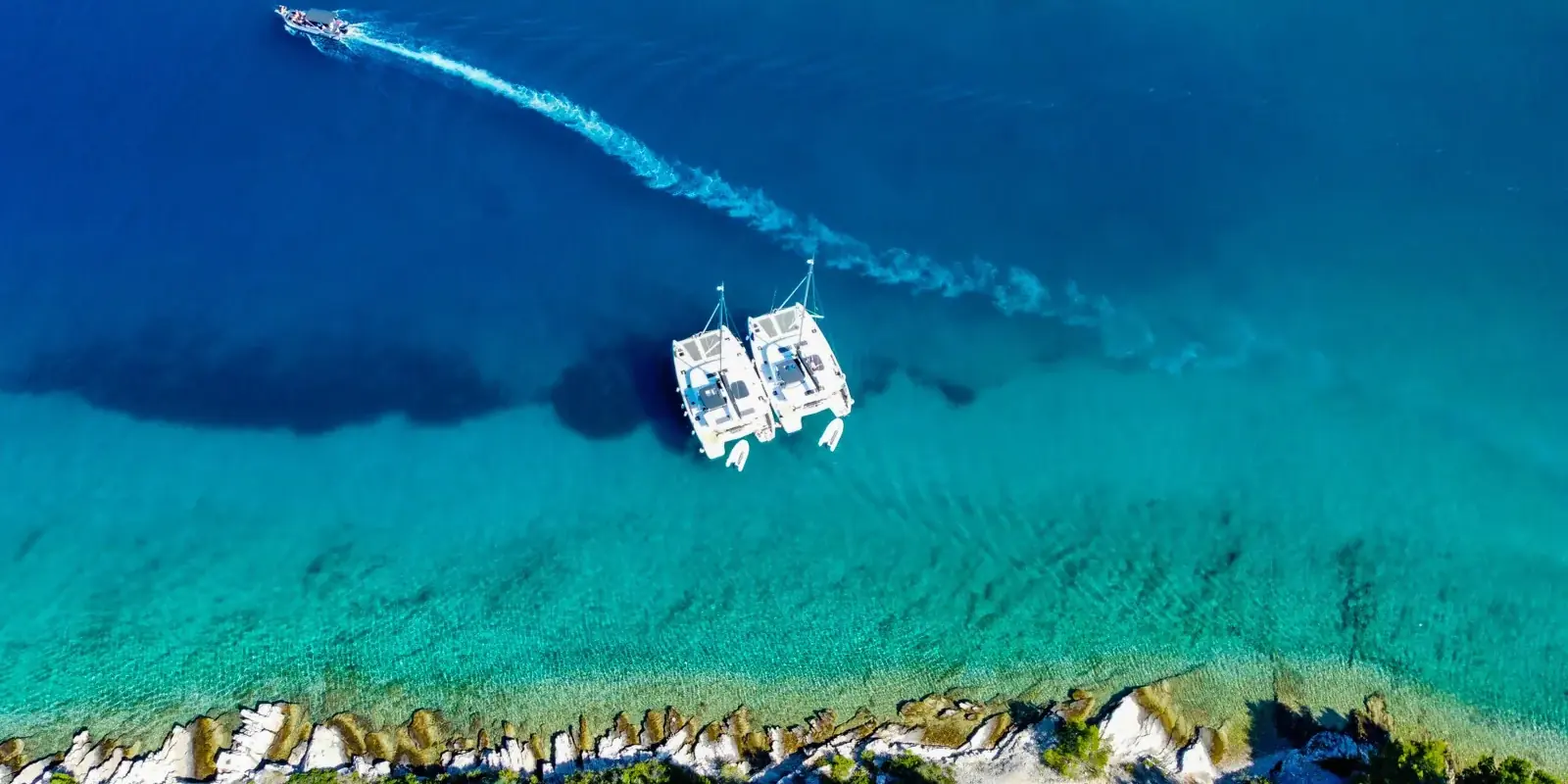 Due catamarani all'ancora sull'acqua turchese lungo una costa rocciosa, visti dall'alto