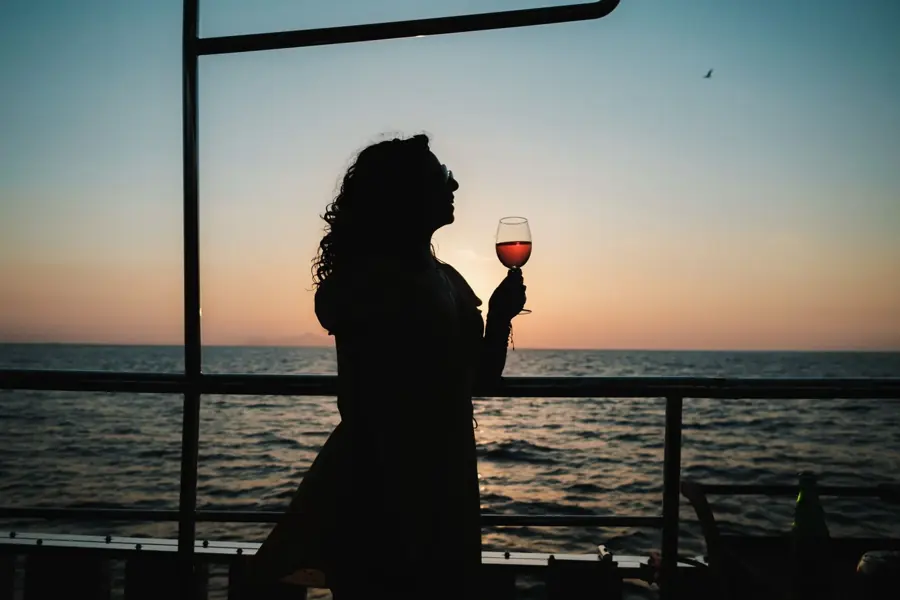 Una figura in controluce con un calice di vino rosso sul ponte di una barca al tramonto